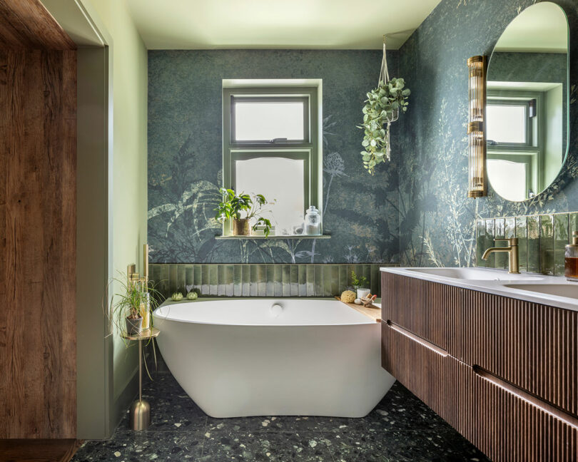 Modern bathroom with a freestanding white bathtub, green botanical wallpaper, round mirror, wooden vanity, plants, and a window letting in natural light.