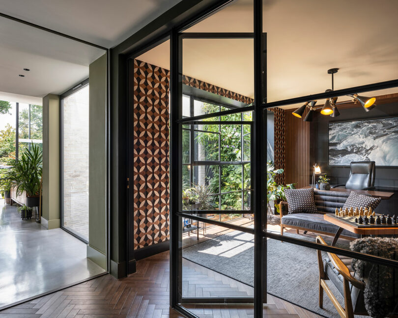 Modern home office with geometric patterned accent wall, glass door, large window, desk, armchairs, and indoor plants visible in adjoining hallway. Natural light fills both spaces.