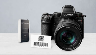 A Lumix S5 II camera, an Opticon barcode scanner, and a card displaying a QR code with a barcode, all placed on a white surface against a light gray background.