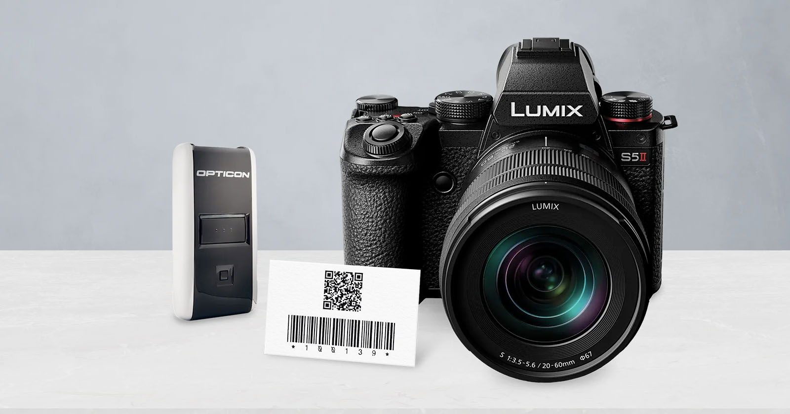 A Lumix S5 II camera, an Opticon barcode scanner, and a card displaying a QR code with a barcode, all placed on a white surface against a light gray background.