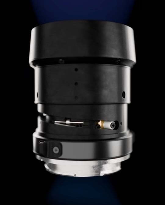 Close-up view of a black camera lens with a metallic mounting ring, shown against a dark background. The lens is facing downward and has visible screws and mechanical details.