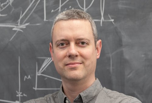 Physics Professor Ryan Patterson stands in front of a chalk board wearing a gray button-down shirt.