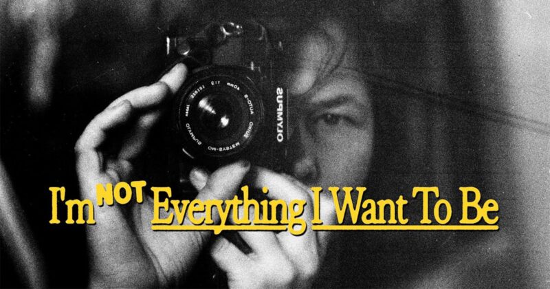 A person holds a camera close to their face, partially obscured in black and white. Yellow text over the image reads, "I'm NOT Everything I Want To Be.