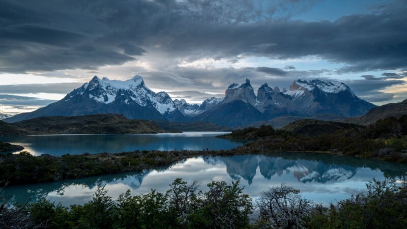 Snow-capped mountains rise above a reflective lake surrounded by rugged hills and green vegetation under a dramatic cloudy sky at sunrise or sunset.
