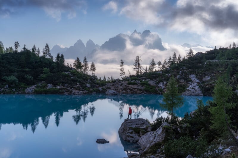 A person in a red jacket stands on a rock by a calm blue lake, surrounded by trees and mountains, with mist and clouds in the background under a dramatic sky.