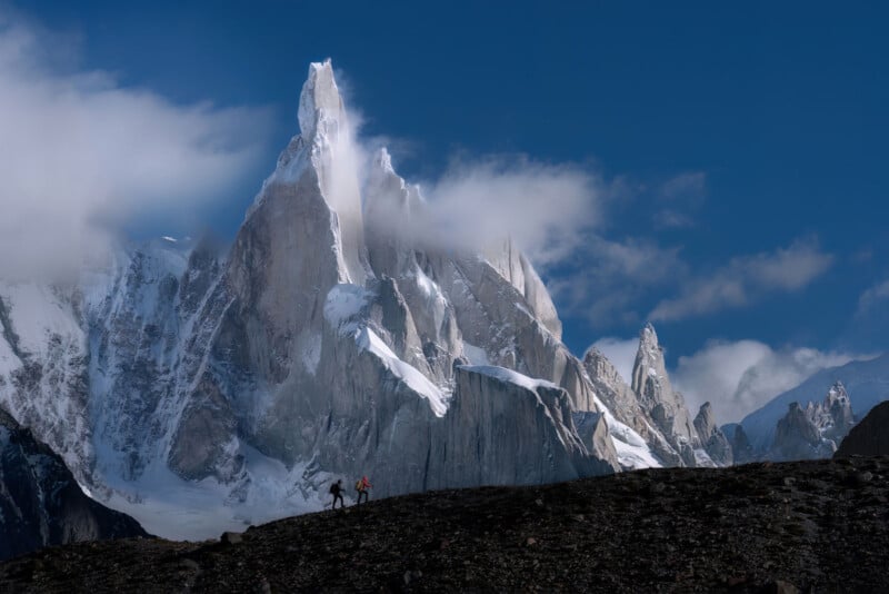 Two climbers trek across rocky terrain in the foreground, with a dramatic, jagged, snow-capped mountain peak rising into the clouds under a vibrant blue sky in the background.