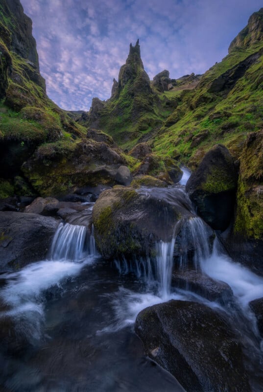 A small waterfall flows over mossy rocks at the base of steep, green cliffs under a blue sky with scattered clouds.