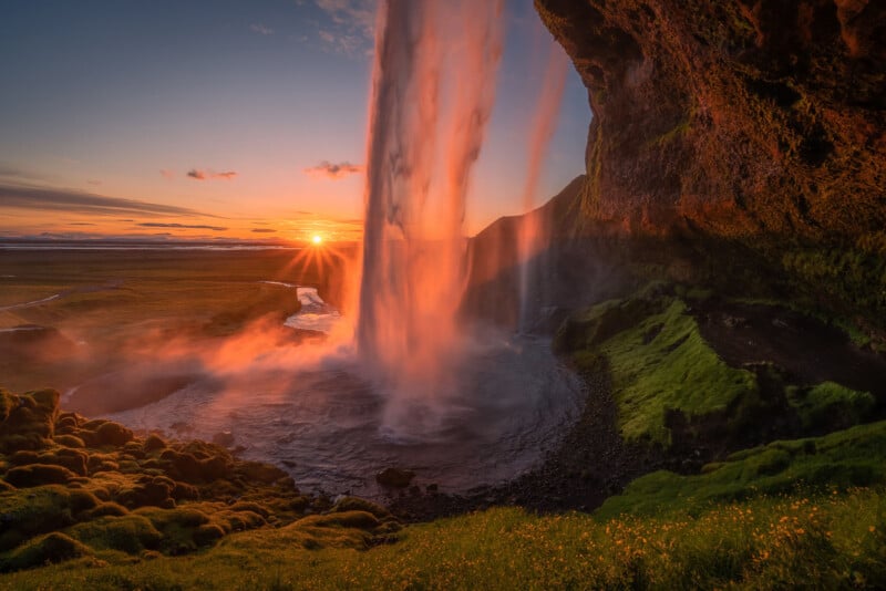 A vibrant sunset illuminates a tall waterfall cascading over a mossy cliff into a pool below, with lush green grass and fields stretching into the distance under a colorful sky.