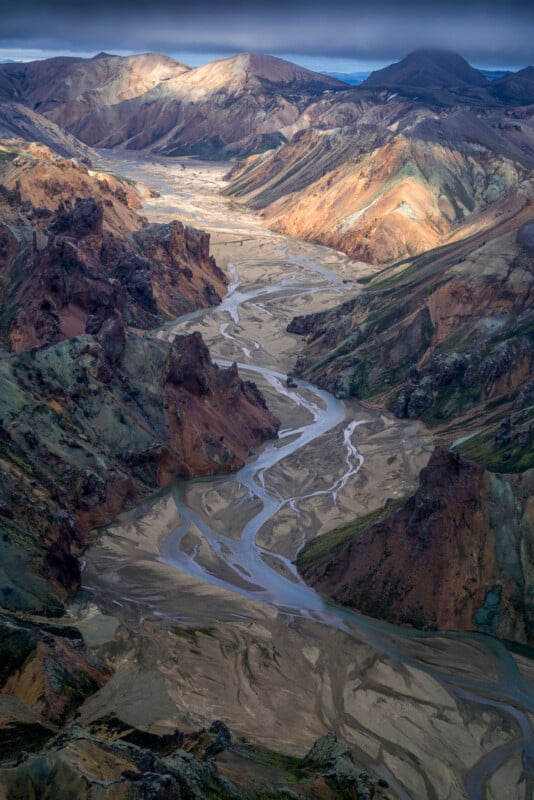 A winding river flows through a wide, rocky valley surrounded by rugged, multicolored mountains under a dramatic, cloudy sky with patches of sunlight highlighting parts of the landscape.