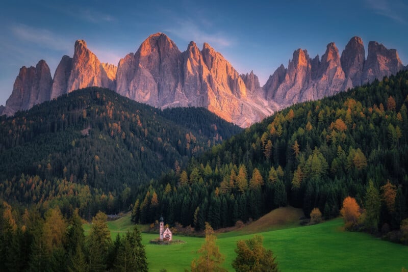 A small chapel sits in a green meadow surrounded by dense forest, with jagged mountain peaks in the background illuminated by warm sunset light.