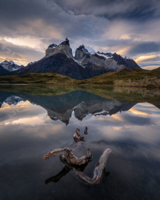 Dramatic mountains are reflected in a still lake at sunset, with driftwood in the water in the foreground and clouds filling the sky.