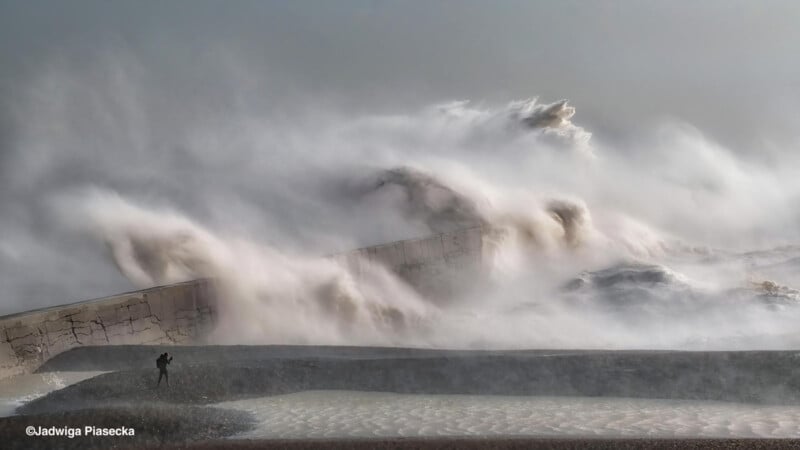 A person stands near a large seawall as massive waves crash over it, creating a dramatic spray of water and mist under a cloudy sky.