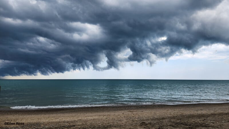 Dramatic dark clouds hang low over a calm blue-green lake, with gentle waves washing onto a sandy beach in the foreground. The scene appears just before a storm.