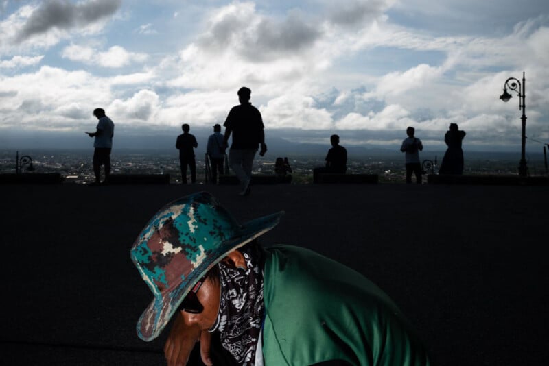 A person in a green camouflage hat and sunglasses is in the foreground, head bowed. Silhouetted figures stand against a cloudy sky and scenic city view in the background. The lighting creates a dramatic contrast.