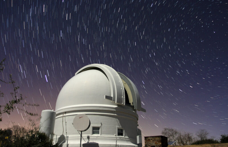 Big, white telescope dome with deep blue sky above it with many stars, appearing as short streaks.