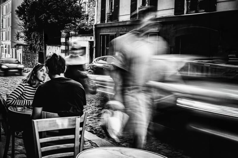 A black and white photo of a couple sitting at an outdoor café table on a cobblestone street, with blurred pedestrians and cars passing by, creating a sense of movement and contrast with the still couple.