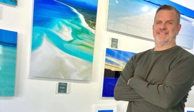 A man with short hair and a beard, wearing a green long-sleeve shirt, stands with arms crossed in front of a wall displaying vibrant landscape photographs, including beaches and ocean scenes.