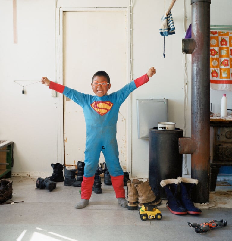 A young child wearing a Superman costume and red glasses stands indoors with arms raised in triumph. The room has boots, a toy truck, a wood stove, and a colorful towel hanging in the background.
