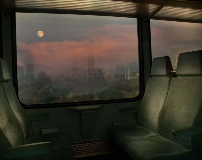 Empty green train seats beside a large window, with a full moon and pink clouds visible over a hazy landscape at dusk outside.
