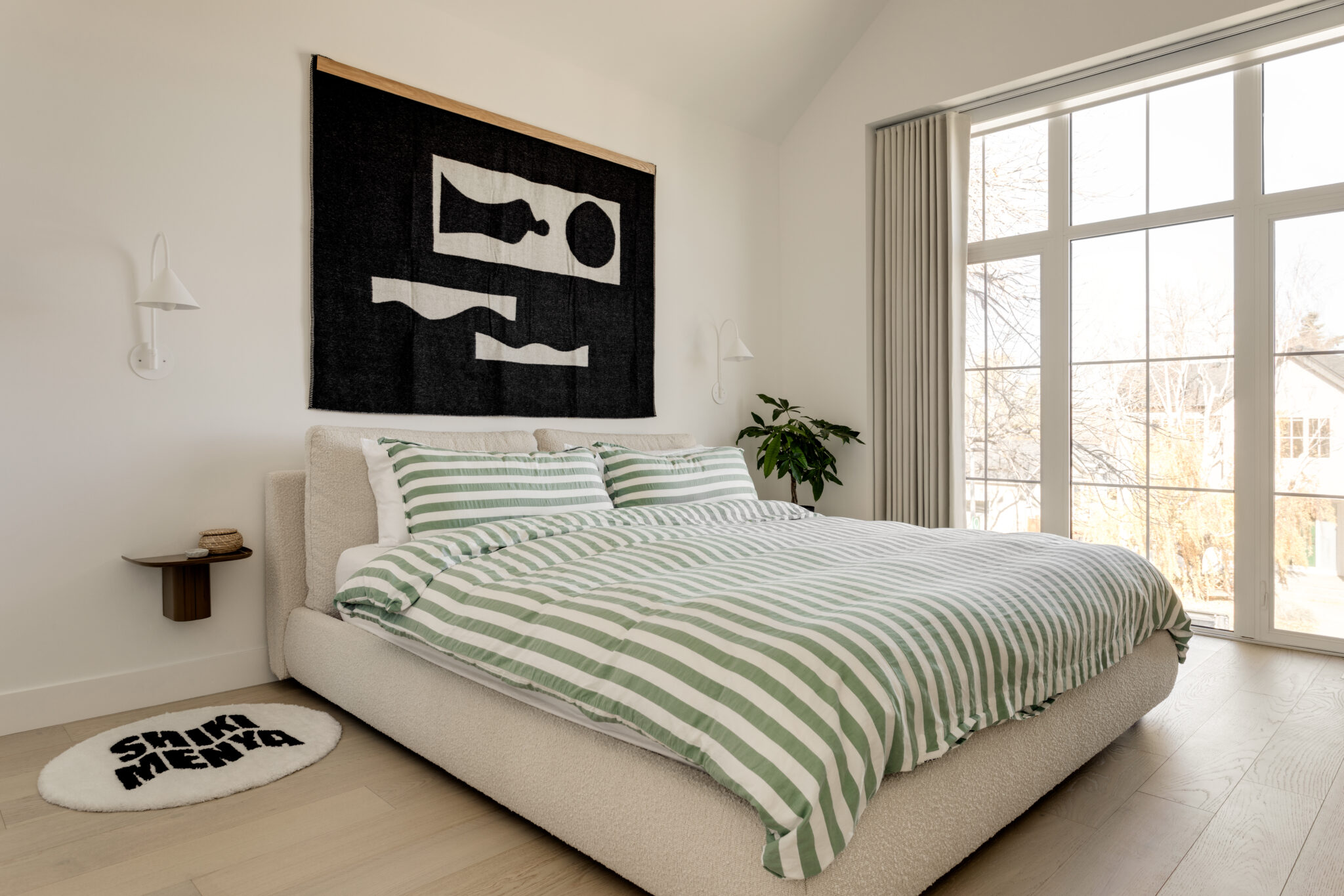 A bedroom with large windows, a bed with green stripped bedding and a large black and white tapestry on the wall.