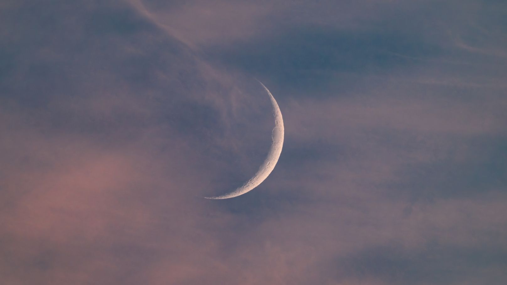 A thin crescent moon is pictured against a darkening blue sky at sunset with a sliver of its right side lit by direct sunlight. Wispy clouds are visible in the foreground.