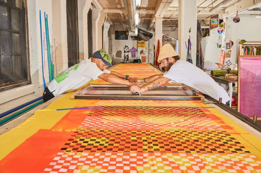 Two artists screenprinting a colorful, graphic print on a large section of orange fabric.