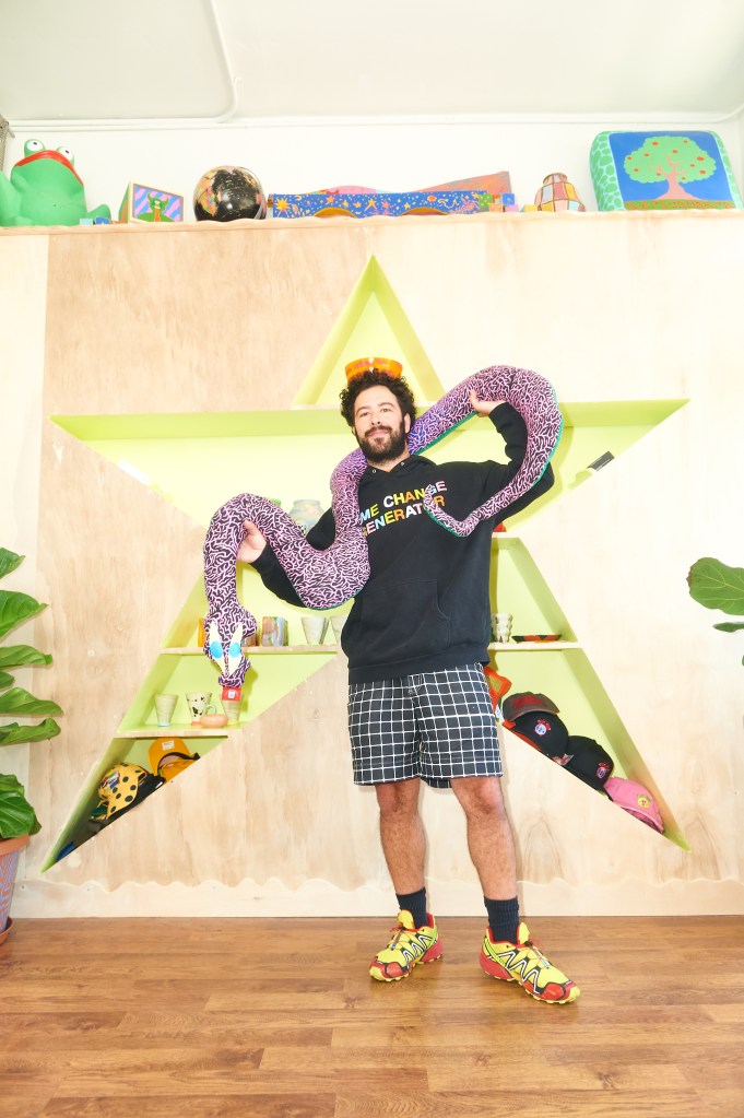 Daniel Bortz standing in front of a star-shaped shelf of ceramics while holding a patchwork snake plush above his head.