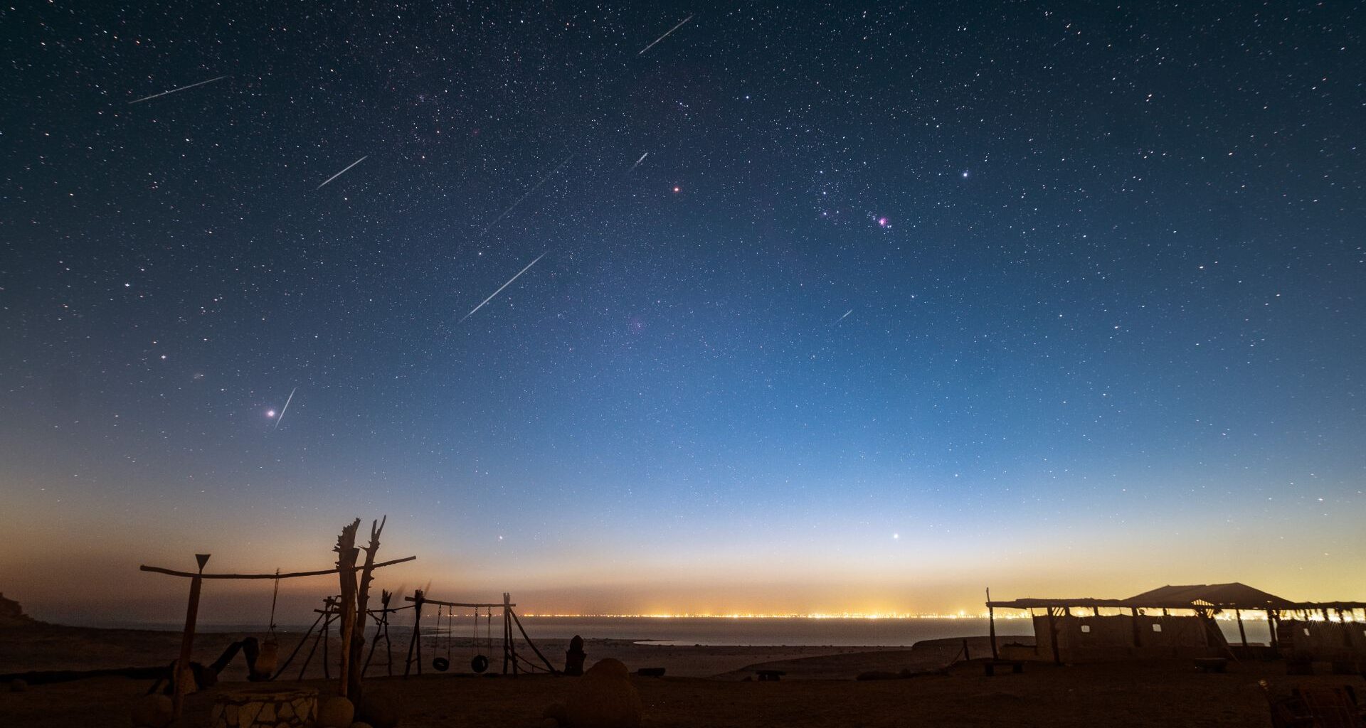 shooting stars are pictured streaking through a starry night sky above a desert. A well and wooden swing set is visible in the foreground, and a glow can be seen creeping into the horizon from a settlement bordering the far side of a lake.