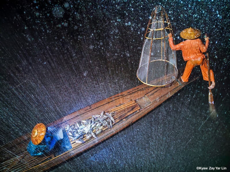 A fisherman wearing an orange outfit and straw hat stands on a wooden boat in heavy rain at night, steering with an oar. Fish are piled on the boat’s deck beside a large fishing net. Water droplets are illuminated by light.