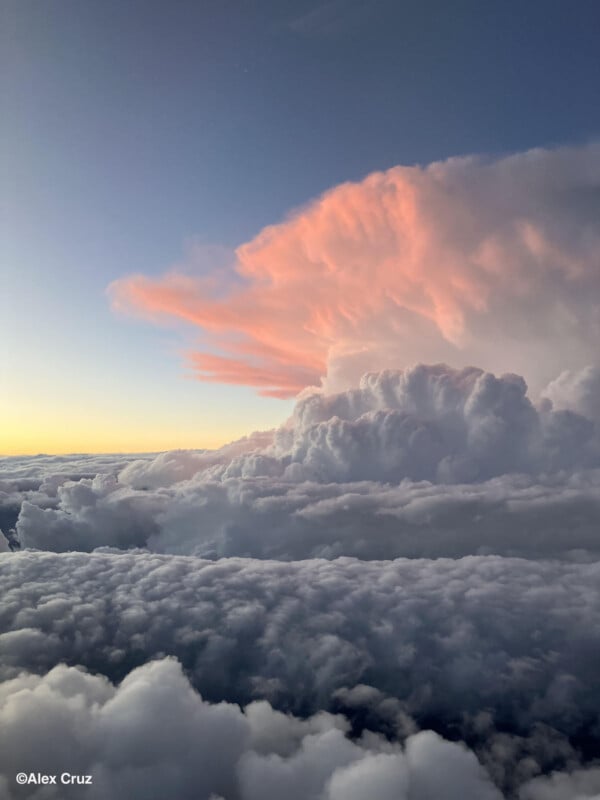 Cloudscape view from above, featuring thick white clouds and a large cumulonimbus cloud lit with soft pink sunlight during sunset or sunrise. Sky fades from blue to yellow. ©Alex Cruz.
