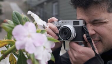 A person holds a camera up to their face, focusing and preparing to take a photo of light pink flowers on a leafy green plant outdoors.