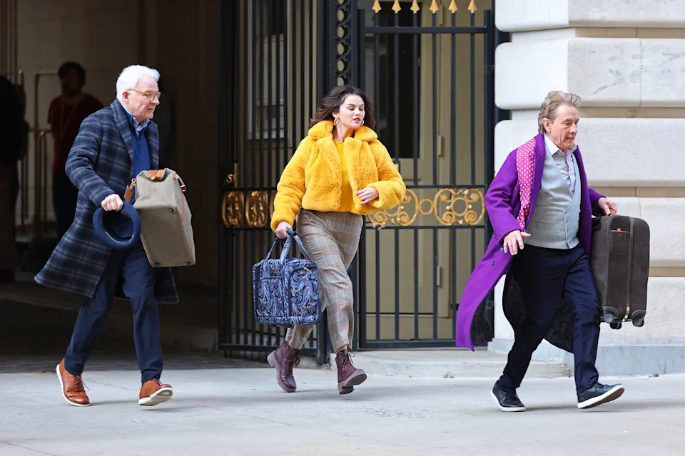Steve Martin, Selena Gomez, and Martin Short carry bags while walking quickly in front of an ornate building entrance