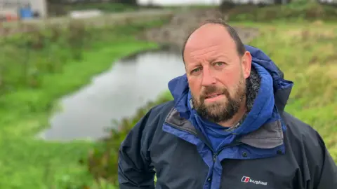 BBC A man stands in front of  a pond on a farm. He is wearing a waterproof coat.   