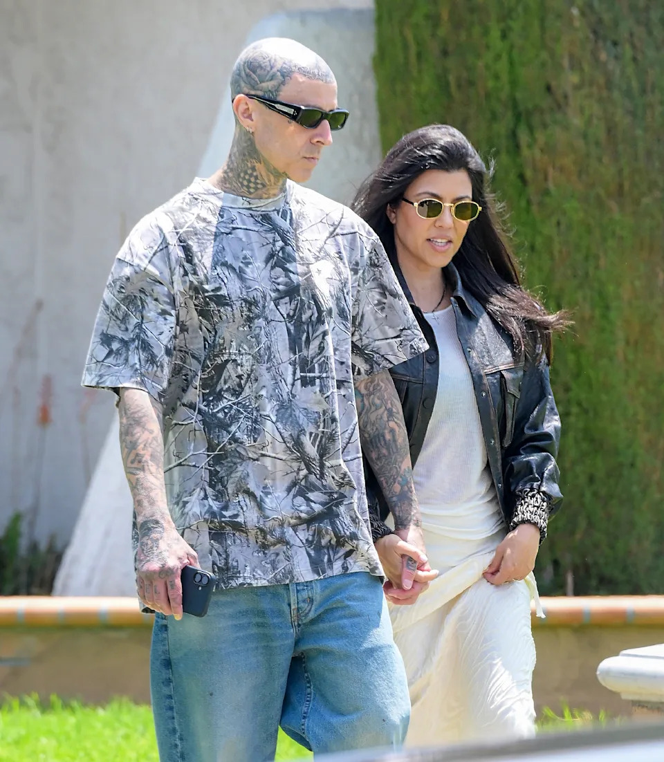Travis Barker is wearing a patterned shirt, and Kourtney Kardashian is wearing a leather jacket and dress, holding hands outdoors, wearing sunglasses