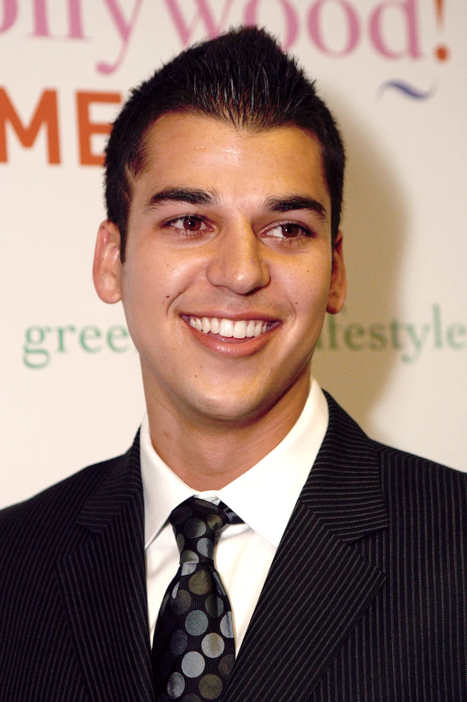 Rob Kardashian is wearing a pinstripe suit and patterned tie smiling at a celebrity event
