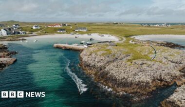 An aerial view of a community in Tiree. There are white-walled houses, a rocky shoreline and two beaches of white sand. The sea is turquoise and a dark green. A kite surfer leaves a frothy wake on the sea's surface as they travel out from a small bay.