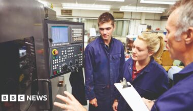 Two young people being shown a complicated piece of machinery by an older man. All three are wearing blue boiler-suits. A small screen with dozens of buttons is attached to the machine.