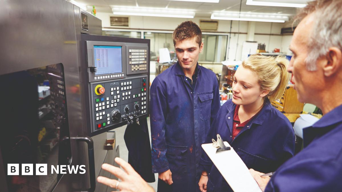 Two young people being shown a complicated piece of machinery by an older man. All three are wearing blue boiler-suits. A small screen with dozens of buttons is attached to the machine.