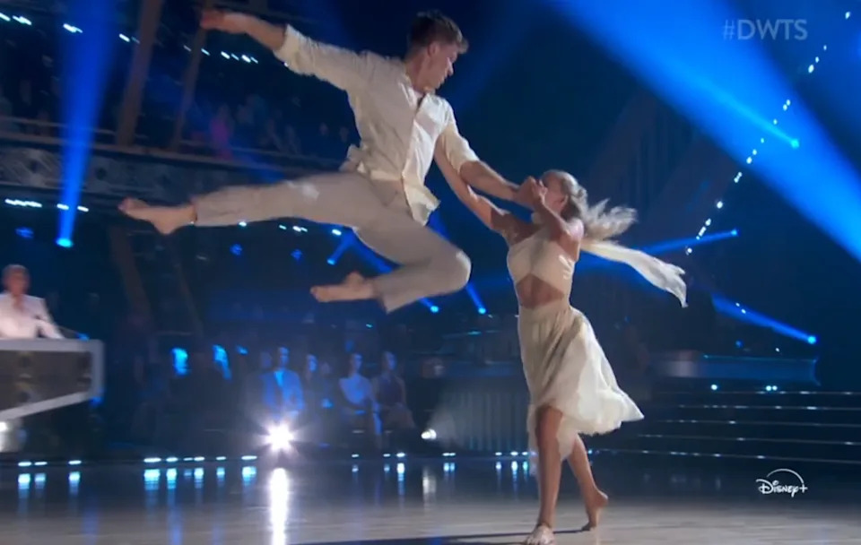 Two dancers perform an energetic routine on "Dancing with the Stars" stage. One leaps dramatically, while the other gracefully extends an arm