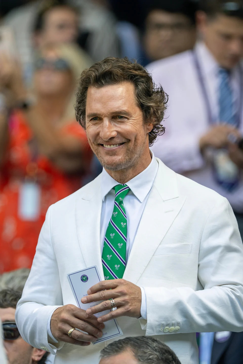 Man in white suit with green patterned tie, holding a pamphlet, smiling at an outdoor event