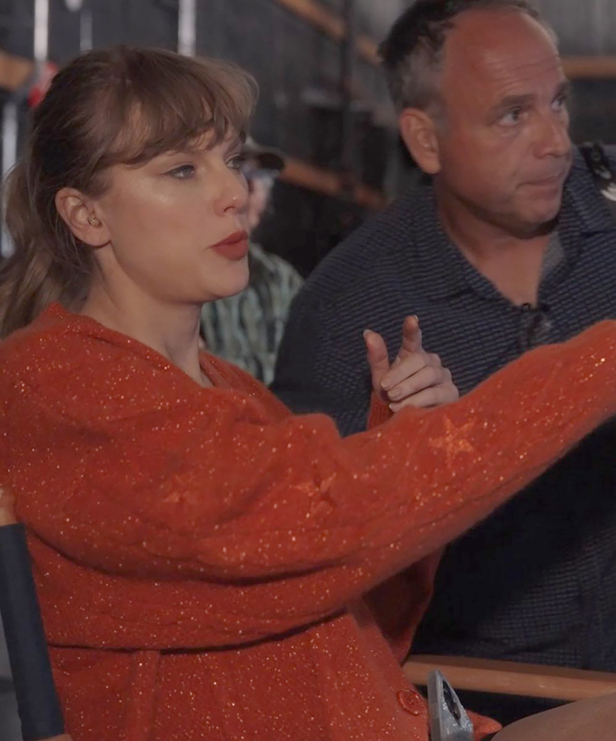 Taylor Swift in a sparkly orange sweater with a man pointing in the background.