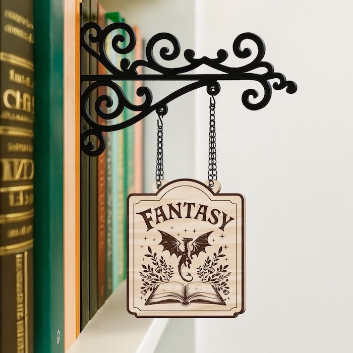 a small wooden tavern sign with the word Fantasy and a dragon hovering over a book on it. the sign is handing from an ornate metal fixture that is places between books ona shelf