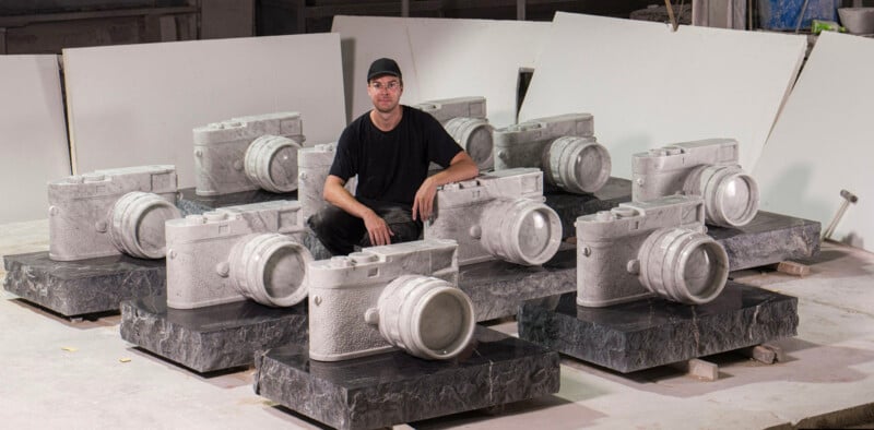 A person wearing a black shirt and cap sits among multiple large stone sculptures shaped like cameras, all placed on stone bases in a workshop setting.