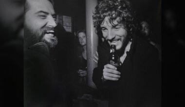 Two men laughing together at a bar, one holding a bottle, with a woman and another person in the background; black and white photo, candid and lively atmosphere.