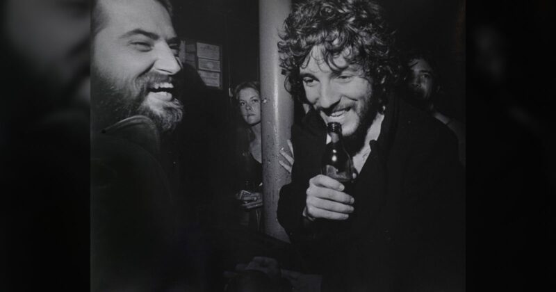 Two men laughing together at a bar, one holding a bottle, with a woman and another person in the background; black and white photo, candid and lively atmosphere.