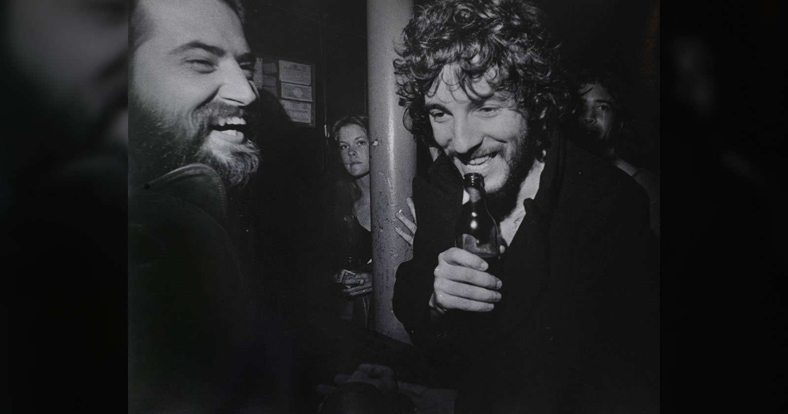 Two men laughing together at a bar, one holding a bottle, with a woman and another person in the background; black and white photo, candid and lively atmosphere.