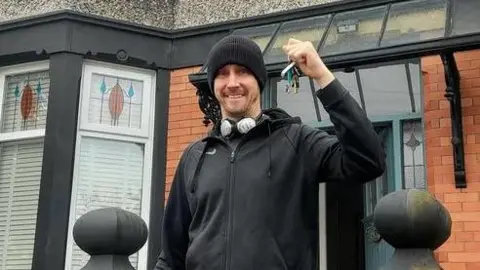 Paul Curtis Street artist Paul Curtis, wearing a black hoodie and beanie hat, smiles as he holds his keys outside his new home