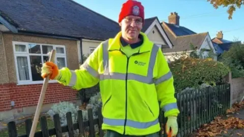 Met Police A smiling man is wearing a yellow fluorescent jacket and is holding a broom handle. He is also wearing a red hat with the Liverpool FC logo on it.
