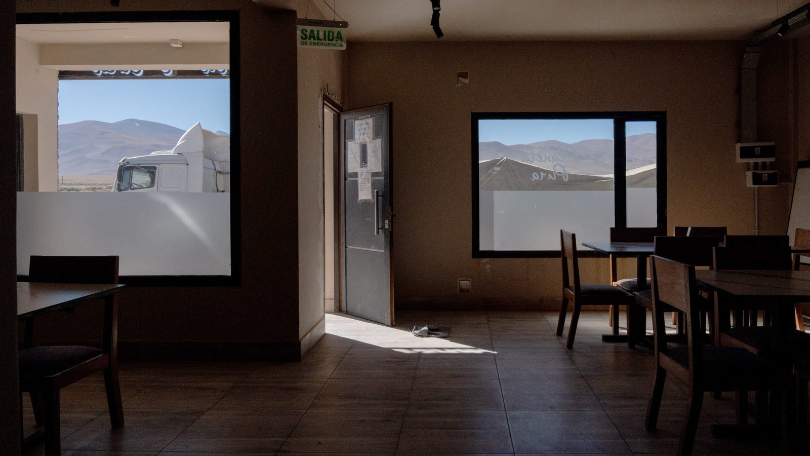 An interior view of a spacious restaurant with wooden tables and chairs, showing an open door leading outside and a large window revealing a mountainous landscape. A truck is partially visible outside the window, with light from the sun creating shadows on the floor.