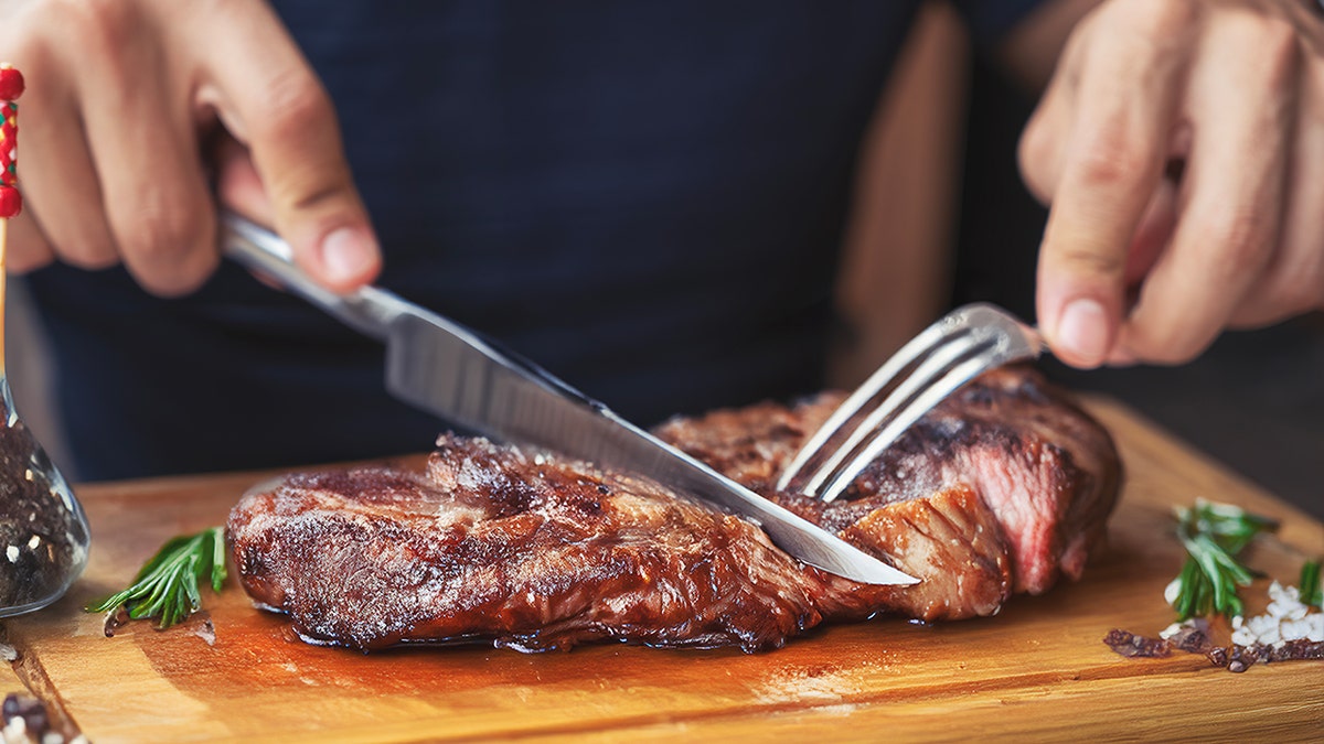 Men's hands holding knife and fork, cutting grilled steak on cutting board with herbs and spices around it.